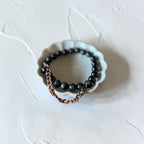 A beaded stretch bracelet with glossy black round beads and a short rose gold chain draped over one side, displayed in a small white scalloped dish on a textured off-white surface.