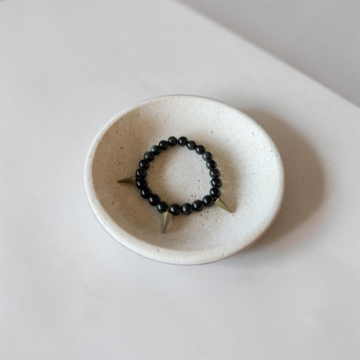 A black beaded stretch bracelet with three metallic spikes, displayed inside a small speckled ceramic dish on a light gray surface.