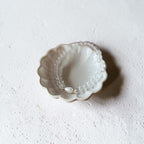 A delicate pearl beaded bracelet with a small gold heart charm rests inside a scalloped, cream-colored ceramic dish on a textured white surface.