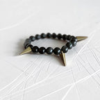 Black bead bracelet with subtle green swirls and brass spikes. Sitting on a white scratch table. 
