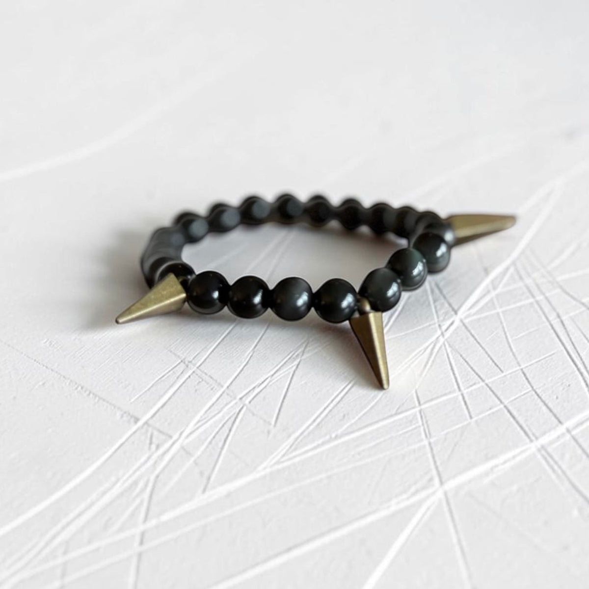 Black bead bracelet with subtle green swirls and brass spikes. Sitting on a white scratch table. 