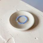 A light blue beaded bracelet with a small white stone accent, displayed on a small round ceramic dish over a textured beige surface.
