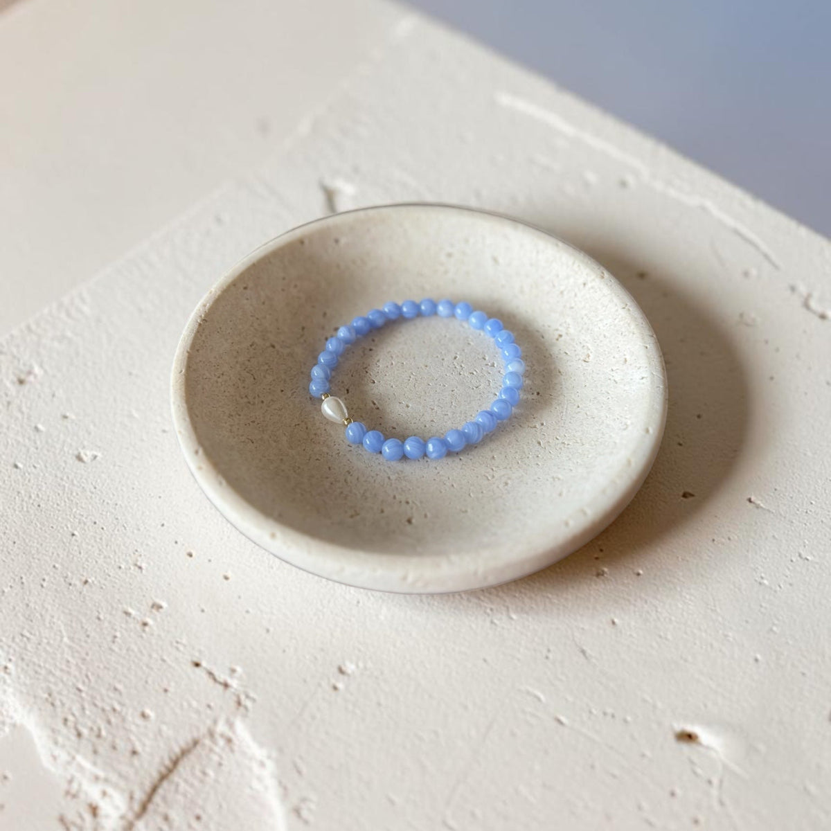 A light blue beaded bracelet with a small white stone accent, displayed on a small round ceramic dish over a textured beige surface.