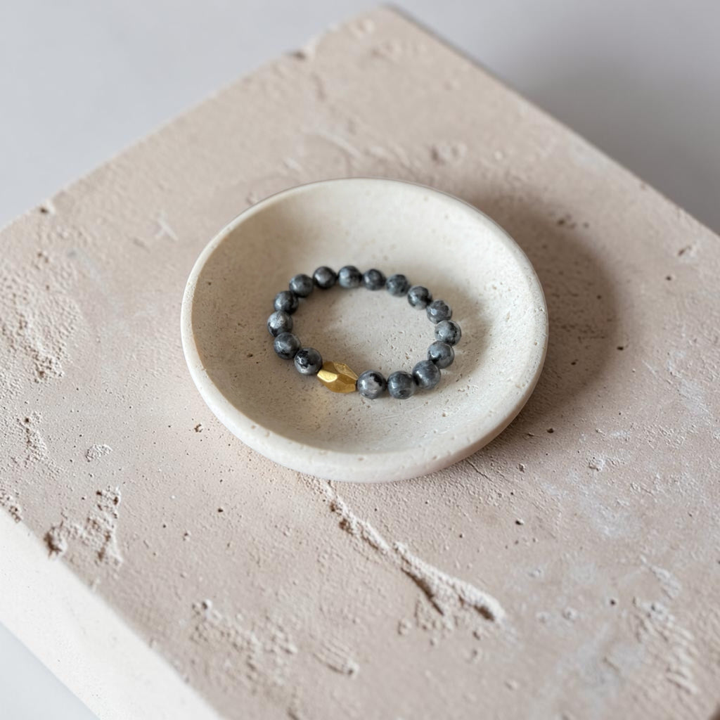 A beaded bracelet with dark gray beads and a small gold accent resting in a small white ceramic dish on a textured beige stone surface.