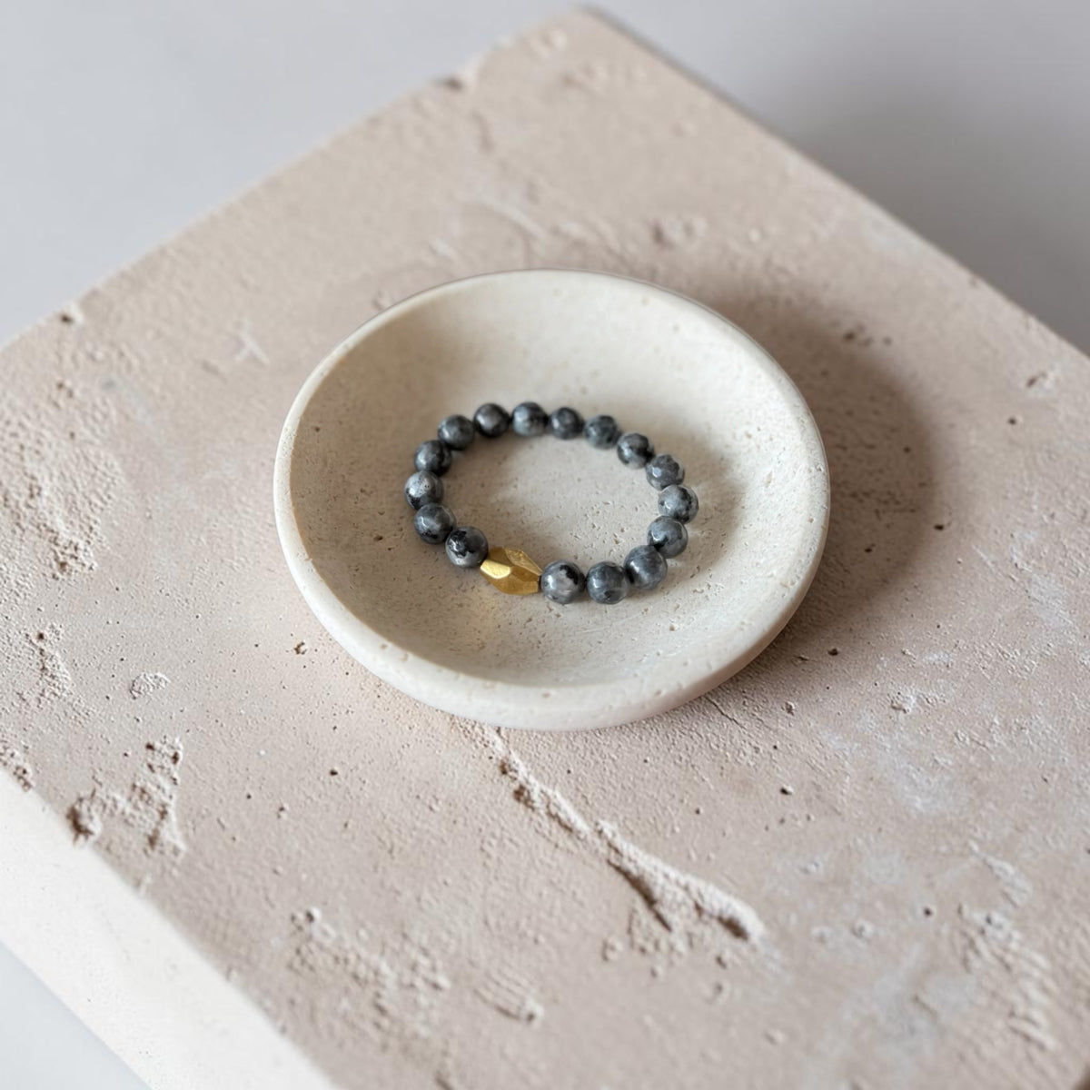A beaded bracelet with dark gray beads and a small gold accent resting in a small white ceramic dish on a textured beige stone surface.