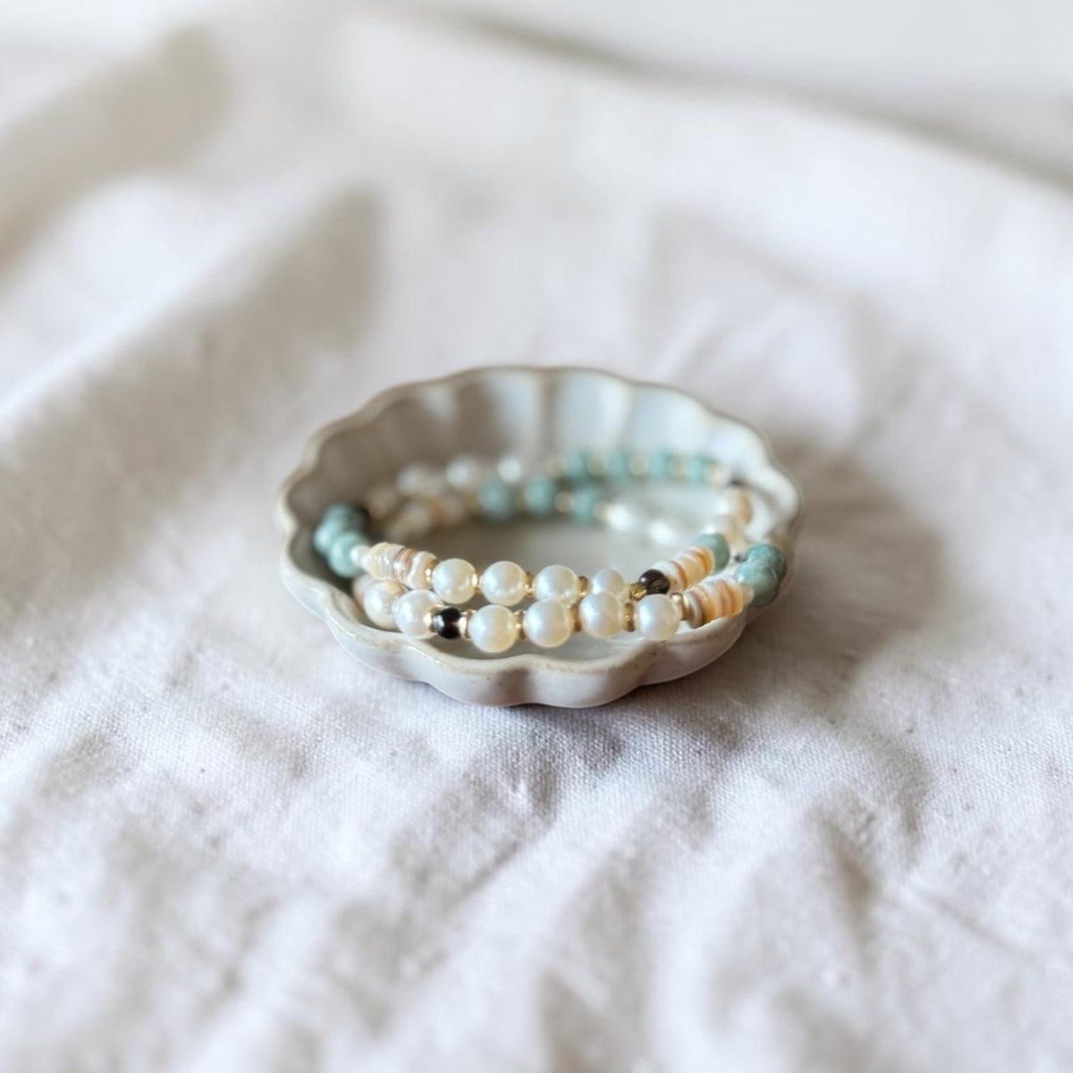 A set of beaded stretch bracelets with white pearls, light blue beads, and small brown accents, displayed in a small scalloped ceramic dish on a soft white fabric background.