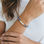 A close-up of a person's wrist wearing a beaded bracelet with blue-gray beads and a central white barrel-shaped bead separated by small gold spacers, against a white shirt background.