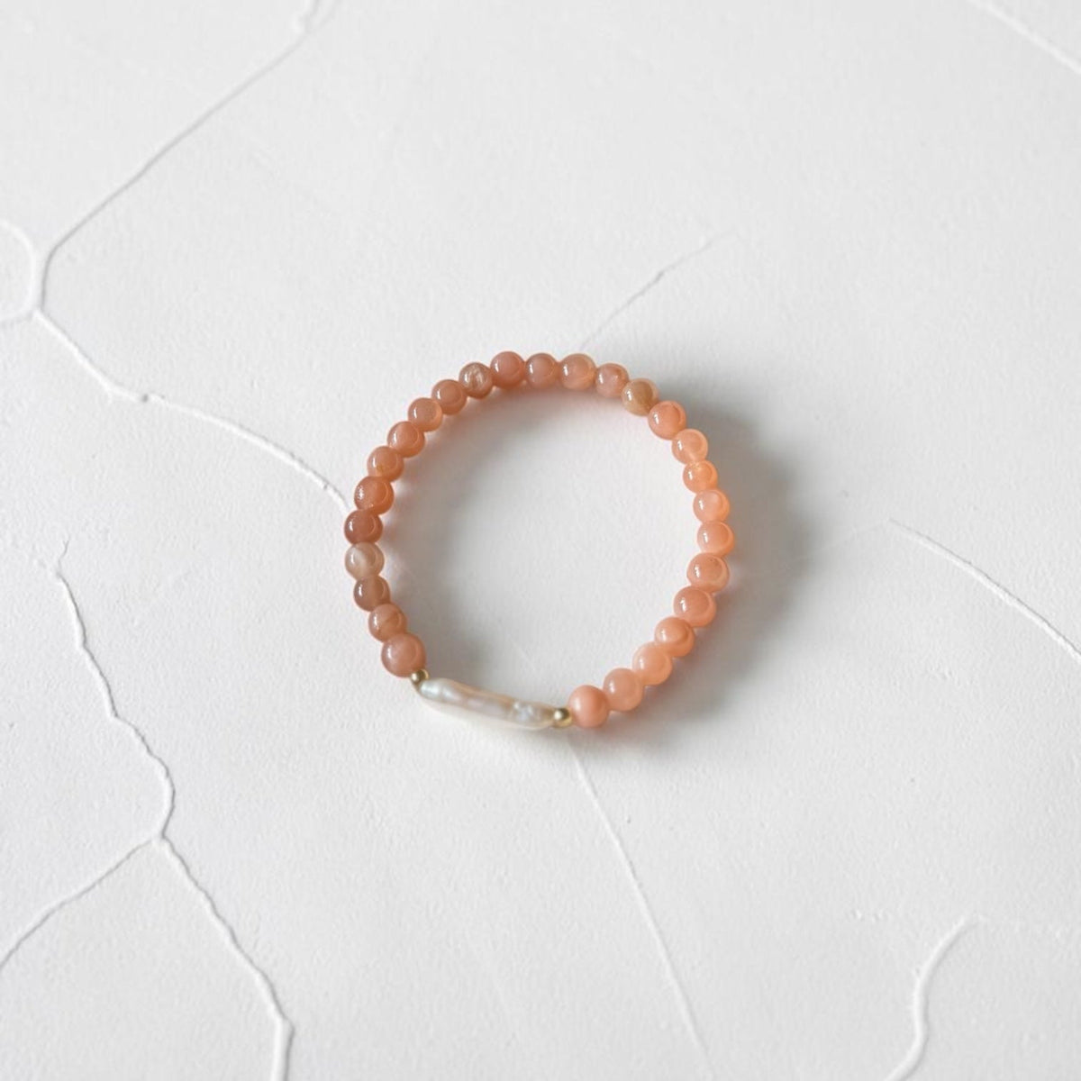A peach-orange beaded stretch bracelet with a small white pearl accent and gold spacer, displayed on a textured white surface.