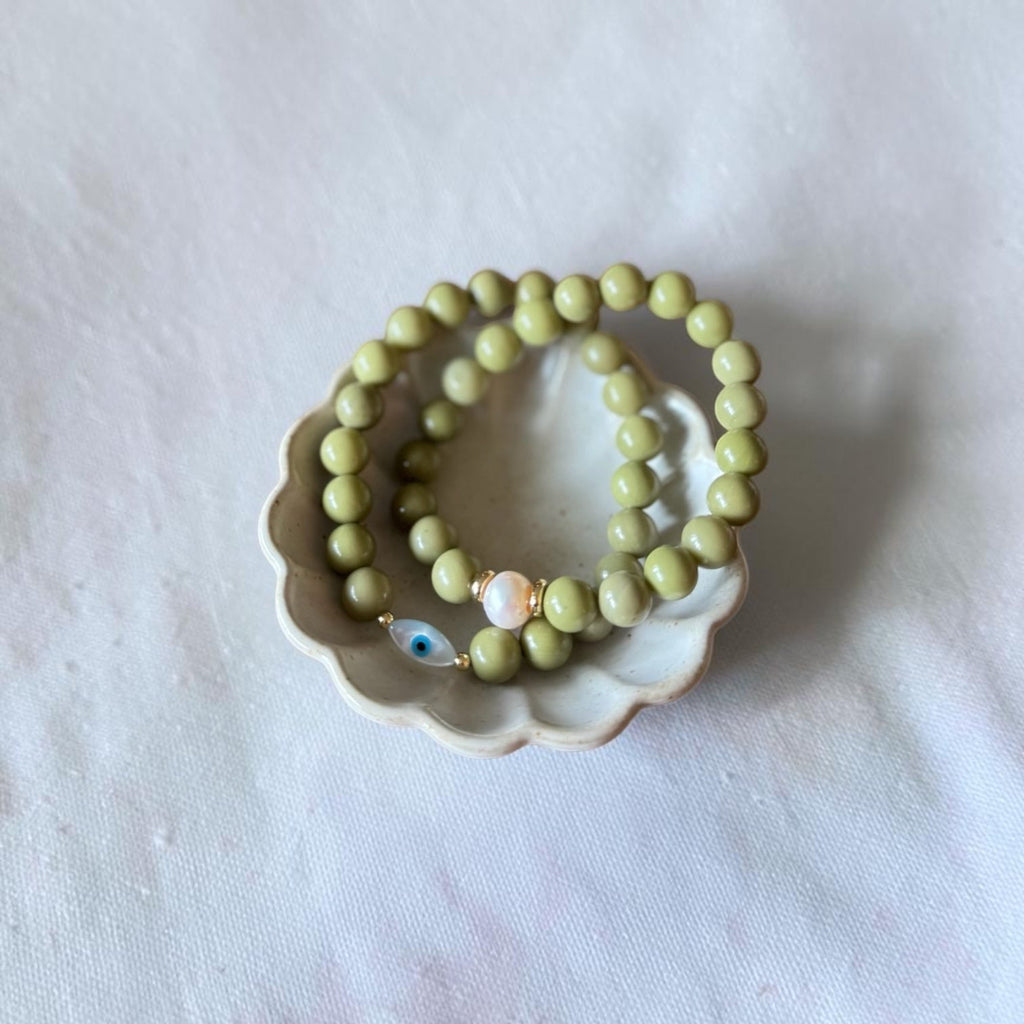 A pair of beaded stretch bracelets in a light olive green color, featuring one bracelet with a blue and white evil eye charm and a gold spacer, displayed on a small beige ceramic dish over a white fabric background.