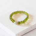 A beaded stretch bracelet made of faceted olive green beads with a single white pearl accent and small gold spacer rings, displayed on a textured white stone block.
