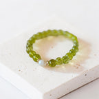 A beaded stretch bracelet made of faceted olive green beads with a single white pearl accent and small gold spacer rings, displayed on a textured white stone block.