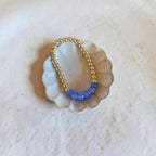 A delicate gold beaded bracelet with a row of small blue beads, displayed on a scalloped off-white dish against a textured light beige background.