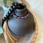 A beaded bracelet with purple amethyst-like beads and small gold spacer beads, draped around a matte black ceramic vase. A black beaded tassel necklace with a tan cord tassel is also placed on top of the vase.