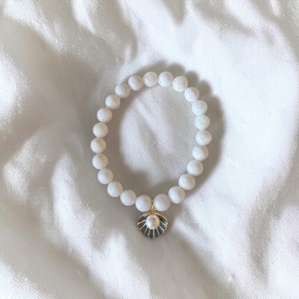 A white beaded bracelet with a small gold-toned shell charm featuring a central pearl, laid flat on a white textured fabric background.