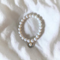 A white beaded bracelet with a small gold-toned shell charm featuring a central pearl, laid flat on a white textured fabric background.