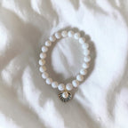 A white beaded bracelet with a small gold-toned shell charm featuring a central pearl, laid flat on a white textured fabric background.