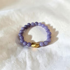 A beaded stretch ring featuring lavender purple round beads and a single faceted gold accent bead, resting on a textured white fabric background.