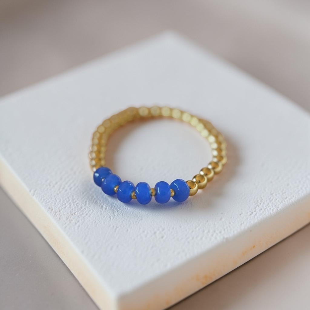 A gold beaded bracelet with five smooth blue beads, displayed on a textured white square block.
