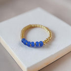 A gold beaded bracelet with five smooth blue beads, displayed on a textured white square block.