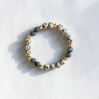 A beaded stretch bracelet featuring white and black speckled beads, dark gray beads, and small gold spacer beads, displayed on a textured white surface with soft shadows.