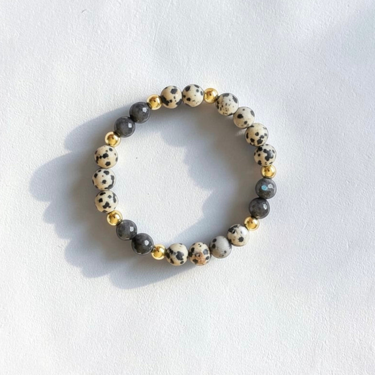 A beaded stretch bracelet featuring white and black speckled beads, dark gray beads, and small gold spacer beads, displayed on a textured white surface with soft shadows.