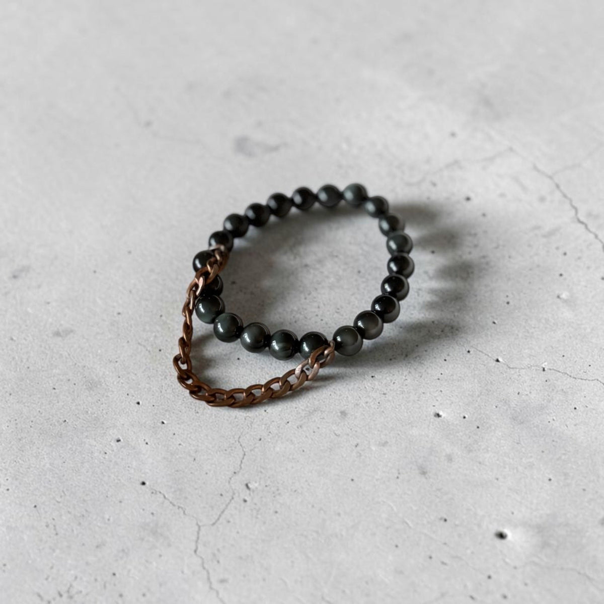 A beaded stretch bracelet featuring dark gray to black round beads and a short copper-toned chain extension, photographed on a light gray textured concrete surface.