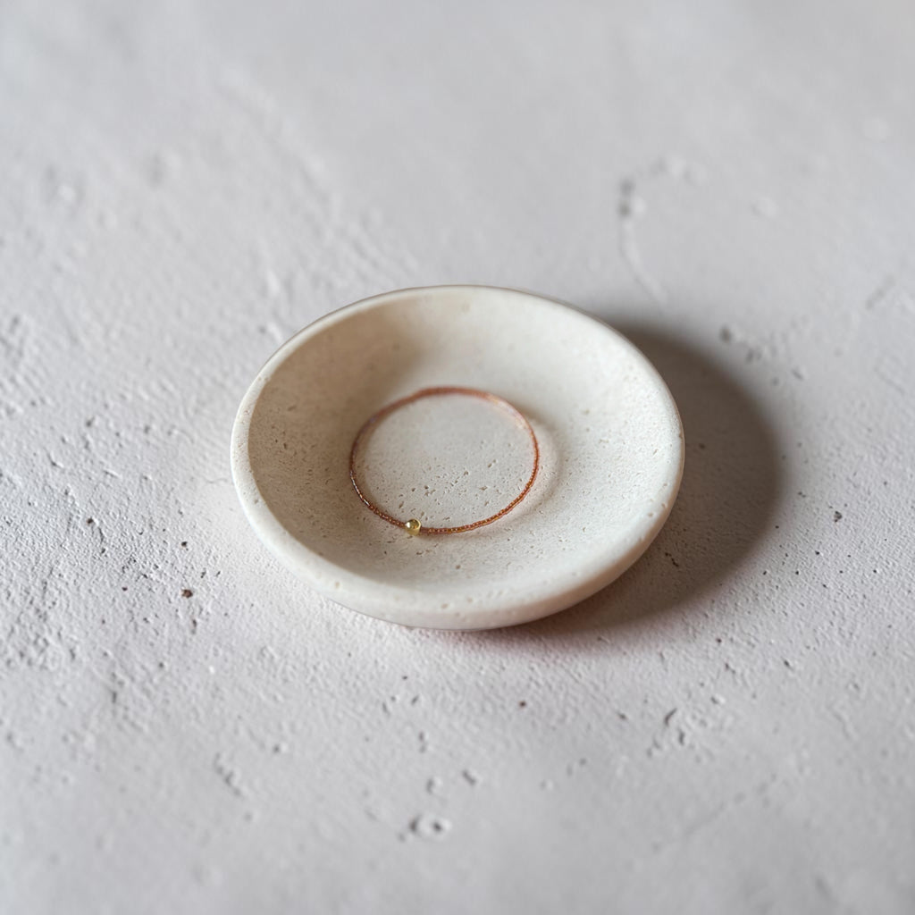 A small round ceramic dish with a speckled off-white glaze, shown on a textured light gray surface with a soft shadow.
