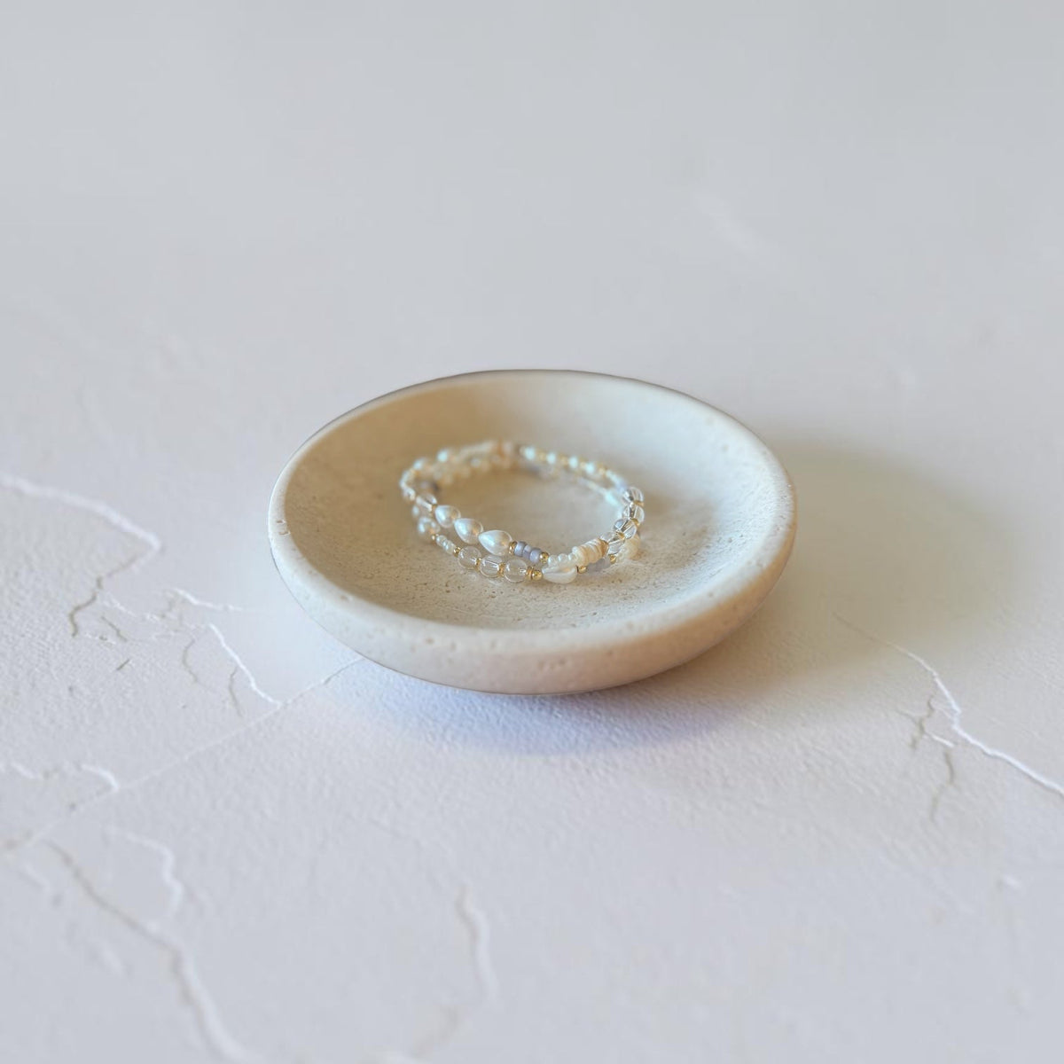 A delicate beaded bracelet with iridescent pearl-like beads sits in a small round ceramic dish on a textured light gray surface.