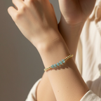 A delicate gold beaded bracelet with five light blue-green round gemstone beads worn on a person's wrist, shown in warm natural sunlight against a soft beige background.