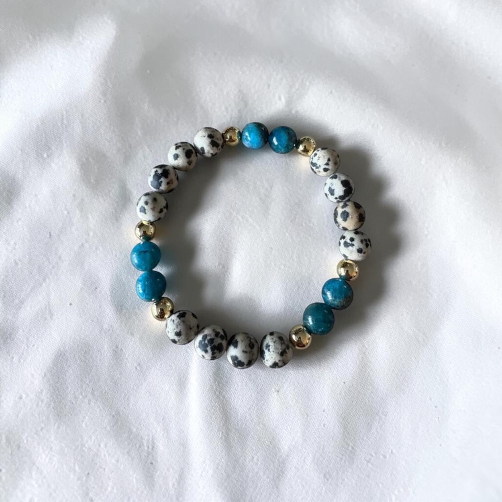 A beaded stretch bracelet featuring white and black spotted round beads, bright blue beads, and small gold spacer beads, displayed on a white fabric background.