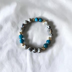 A beaded stretch bracelet featuring white and black spotted round beads, bright blue beads, and small gold spacer beads, displayed on a white fabric background.