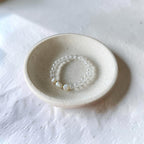 A delicate beaded bracelet with clear round beads and three small white pearl-like beads, displayed inside a small speckled ceramic dish on a textured white surface.