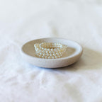 A stack of pearl-bead bracelets displayed in a small round ceramic dish on a soft white fabric background.