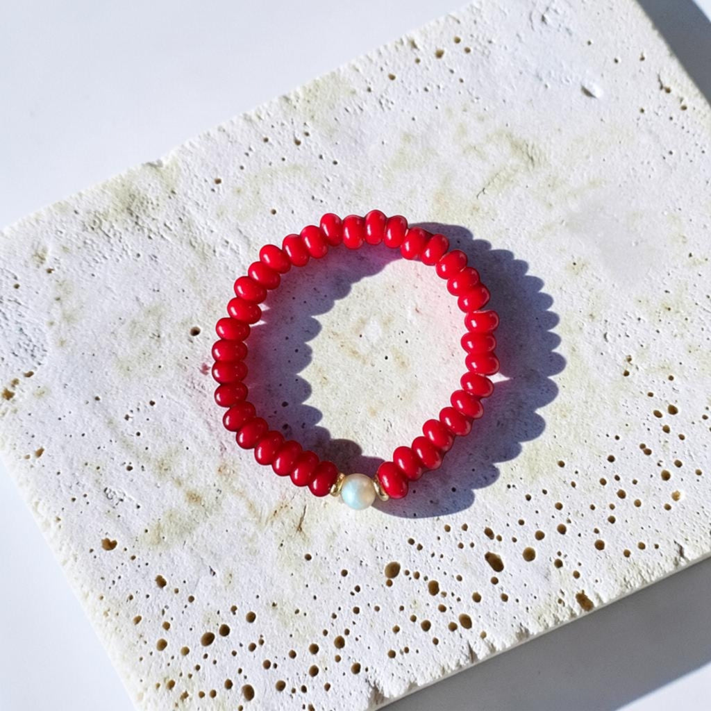 A red beaded stretch bracelet with a small white pearl accent bead, displayed on a textured white stone tile under bright sunlight.