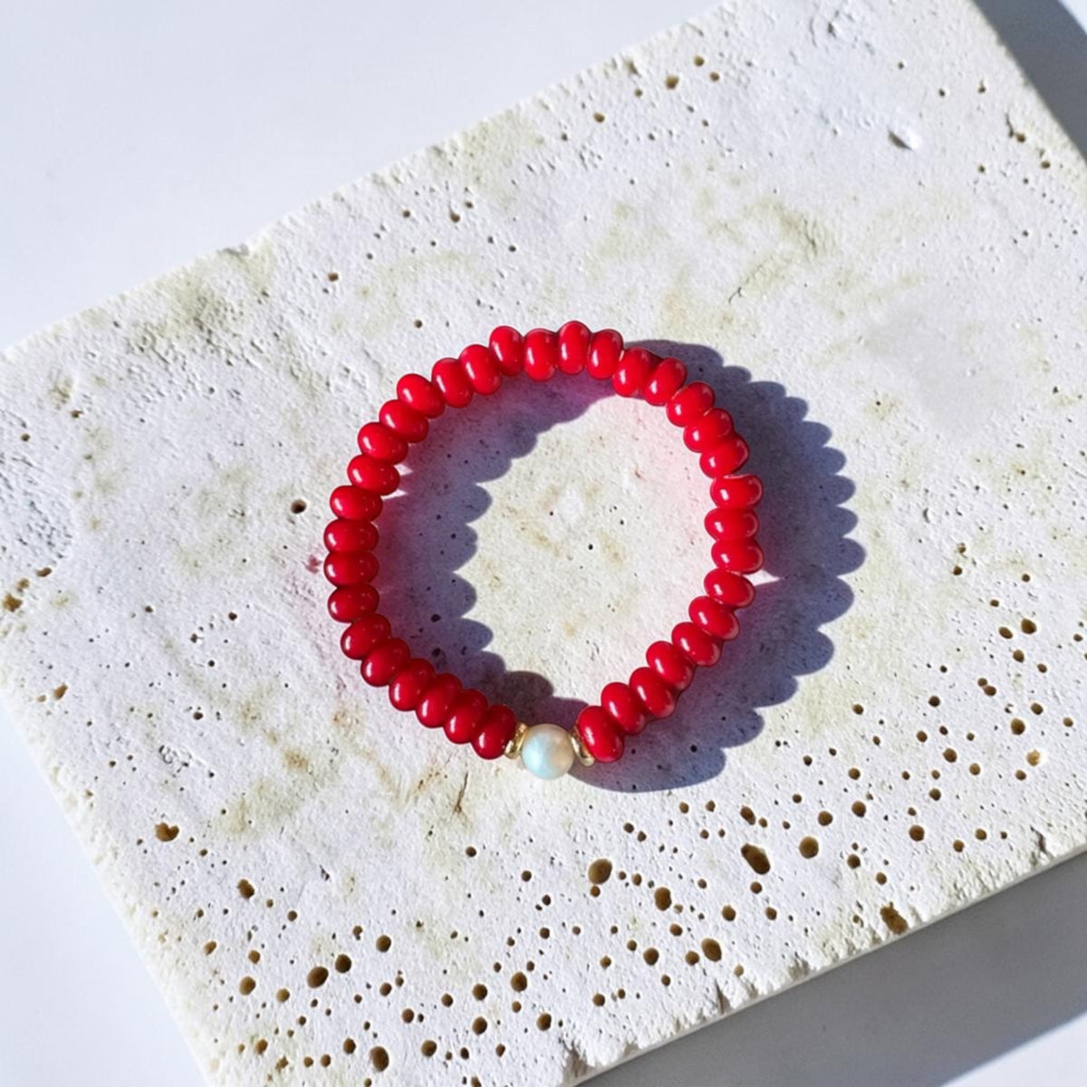 A red beaded stretch bracelet with a small white pearl accent bead, displayed on a textured white stone tile under bright sunlight.