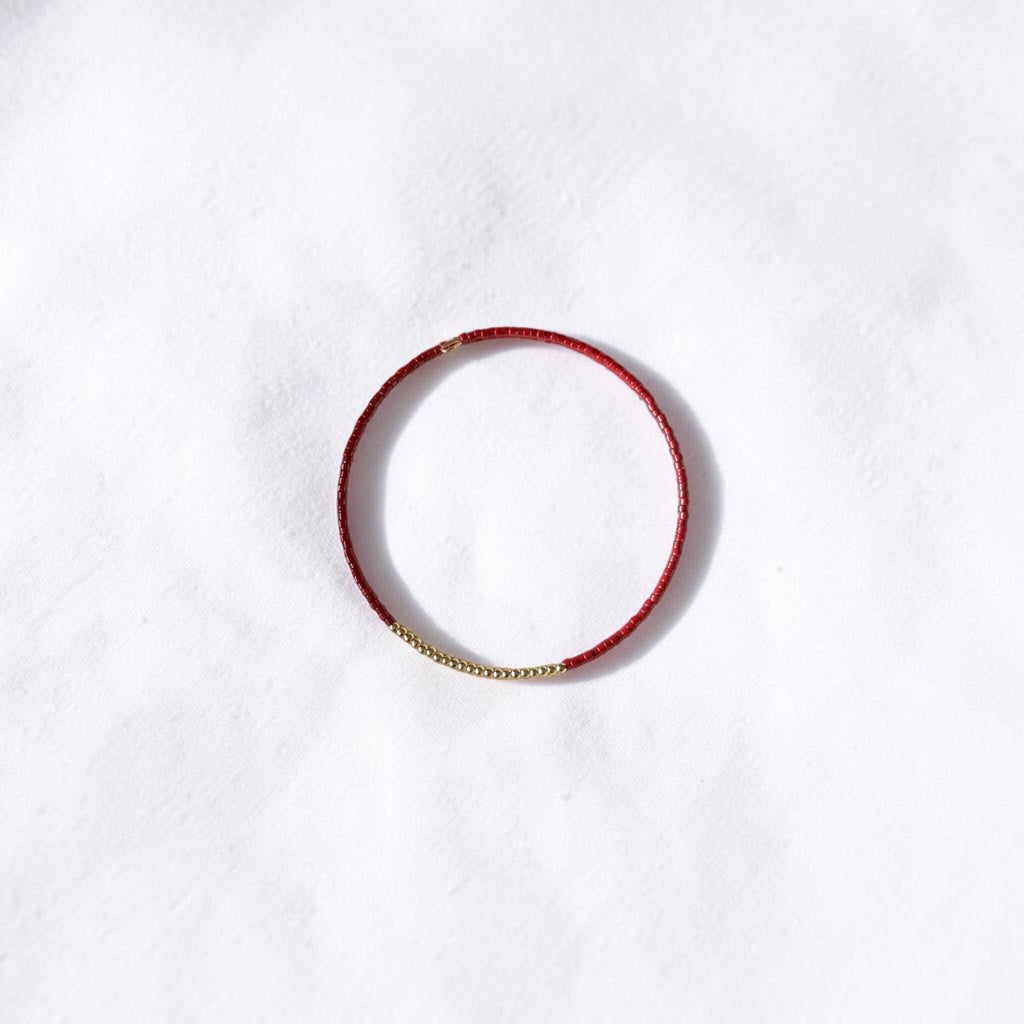 A delicate red beaded bracelet with a small gold chain accent on one side, displayed on a textured white background.
