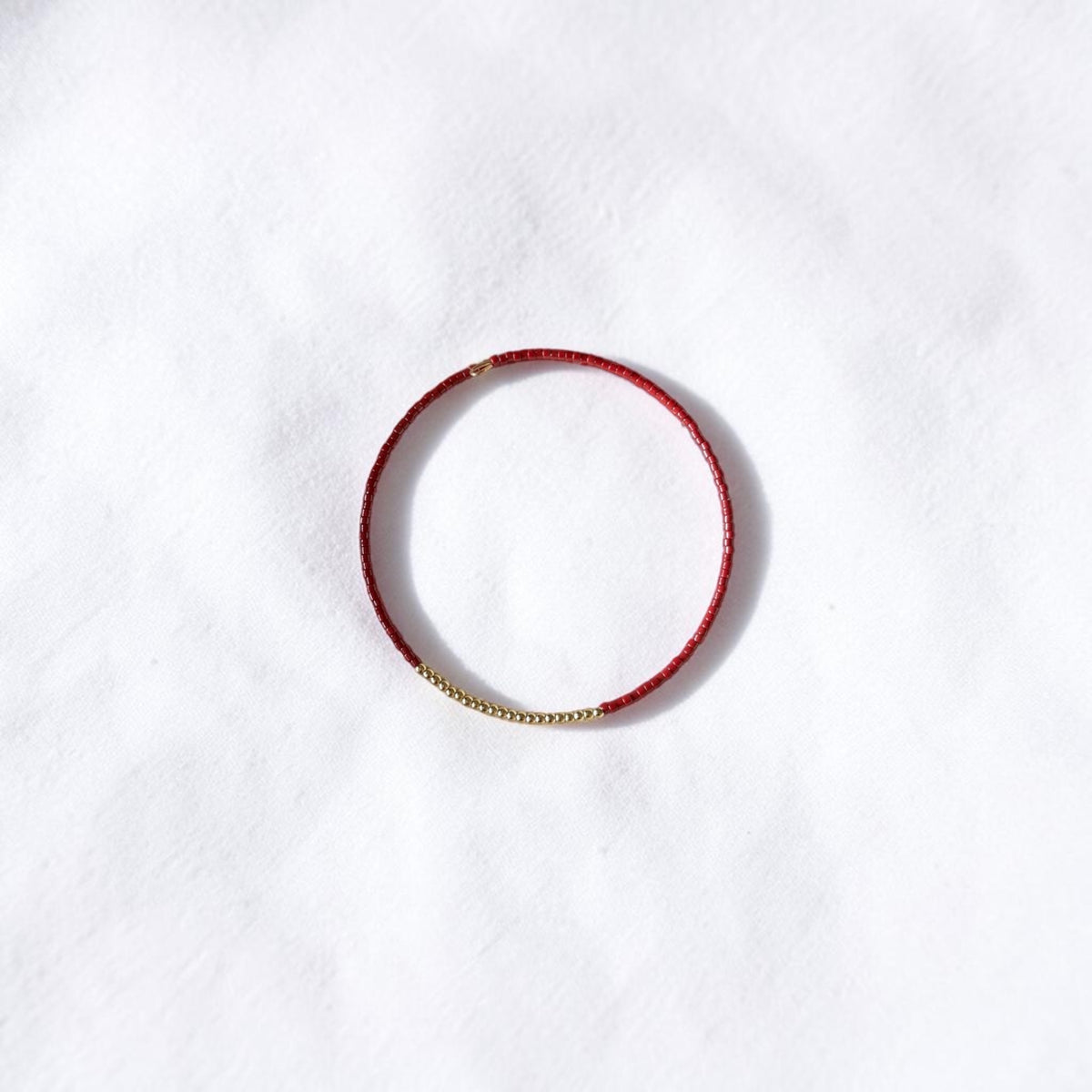 A delicate red beaded bracelet with a small gold chain accent on one side, displayed on a textured white background.