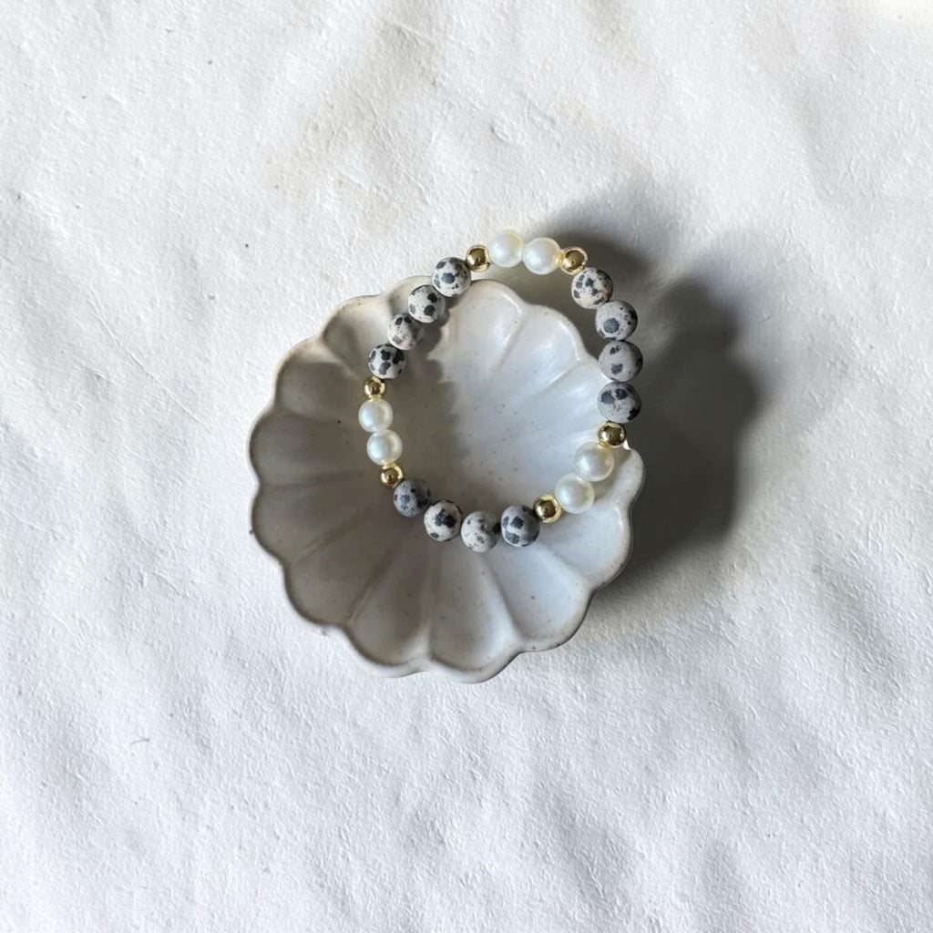 A beaded stretch bracelet with white pearl-like beads, gray and white marbled beads, and small gold spacer beads, displayed on a scalloped white ceramic dish over a textured white fabric background.