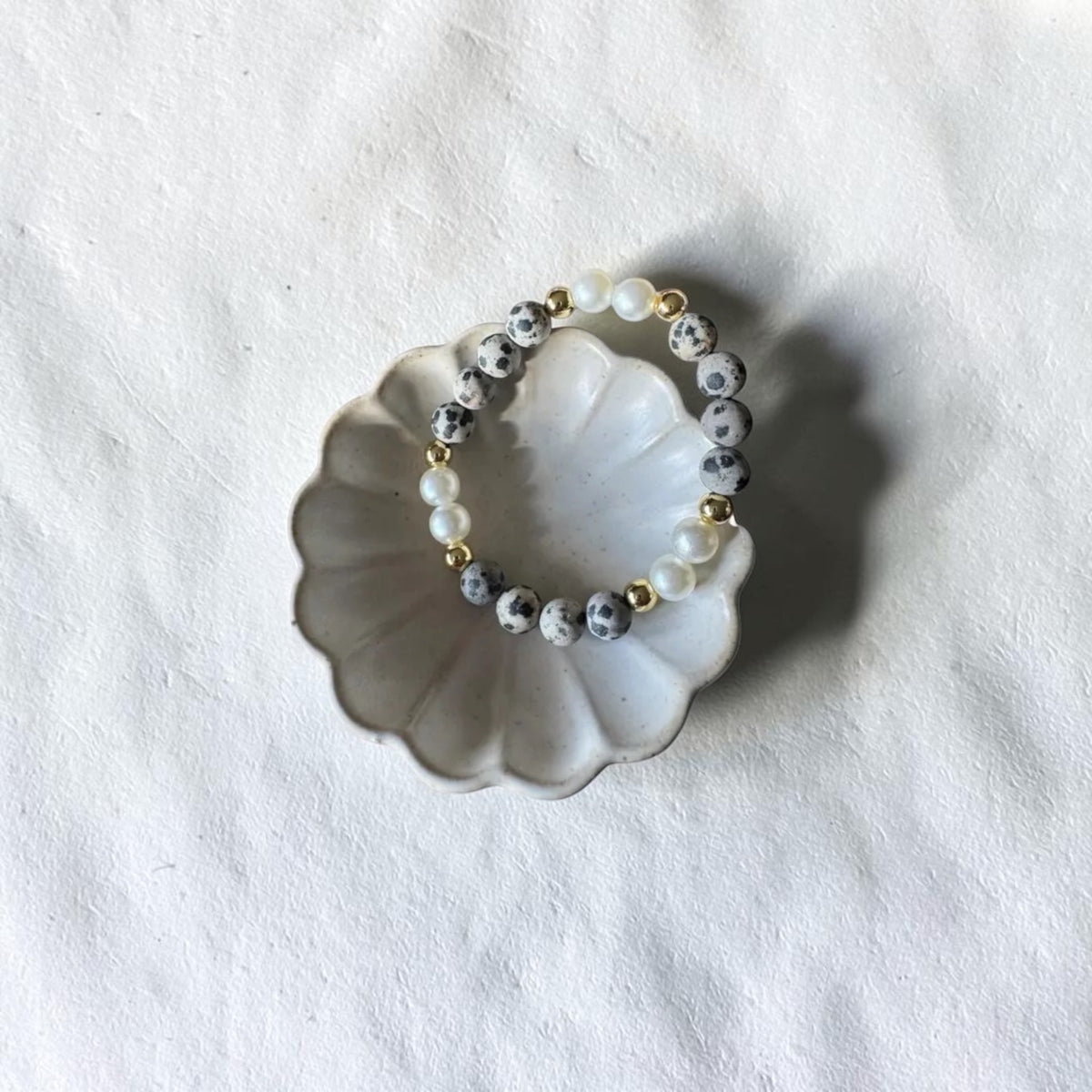 A beaded stretch bracelet with white pearl-like beads, gray and white marbled beads, and small gold spacer beads, displayed on a scalloped white ceramic dish over a textured white fabric background.