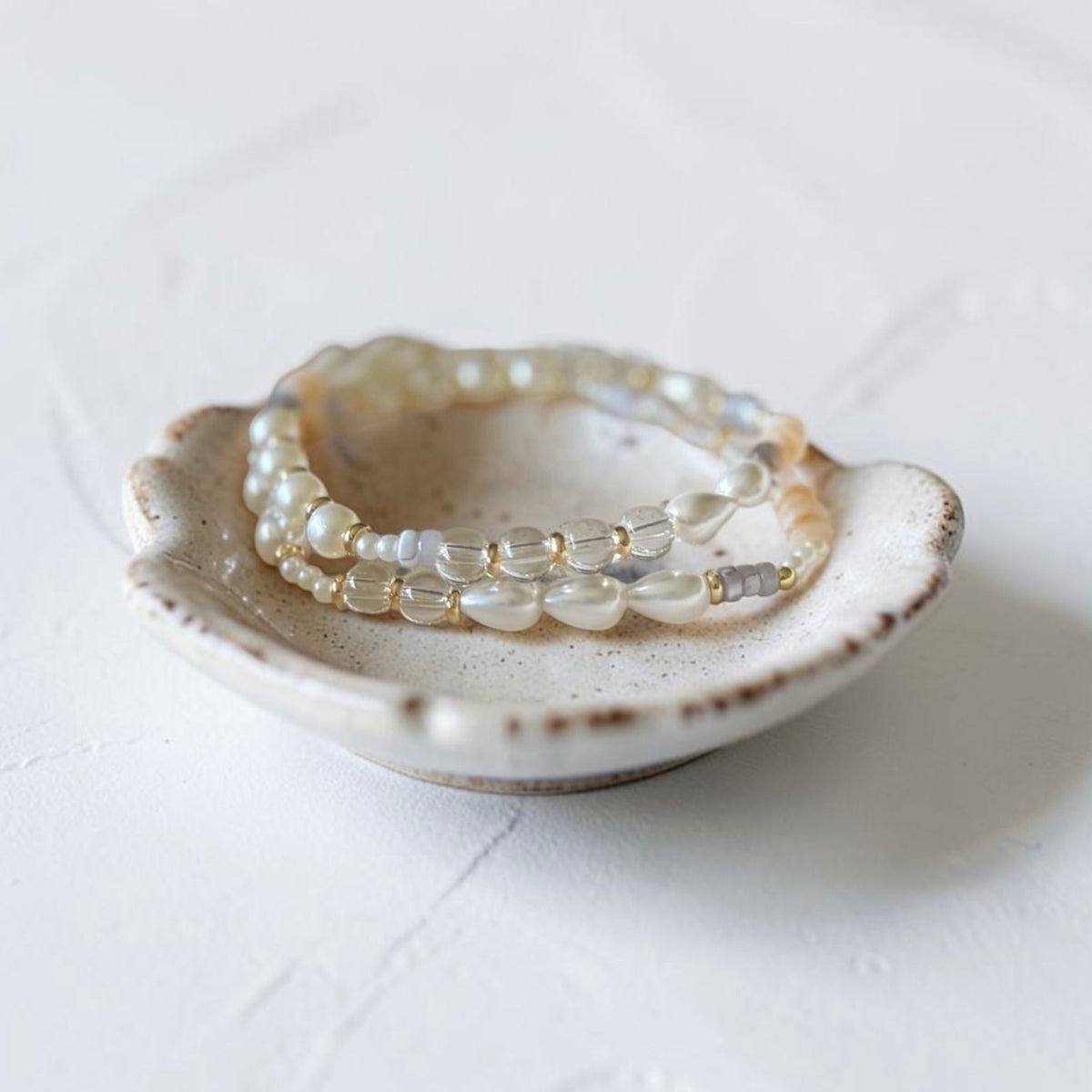 A pair of beaded stretch bracelets with translucent white and pale gold beads, displayed inside a small, rustic ceramic dish on a textured white surface.