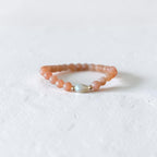 A beaded stretch bracelet featuring peachy-orange stone beads and a central iridescent white bead, displayed on a textured white surface.