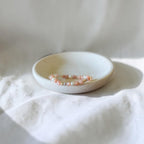 A delicate beaded bracelet in soft pink and white tones rests inside a small, shallow, speckled ceramic dish on draped white fabric.