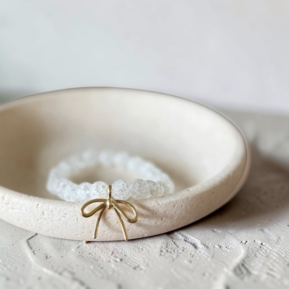 A white beaded bracelet with a small gold bow charm, displayed inside a shallow cream-colored ceramic dish.