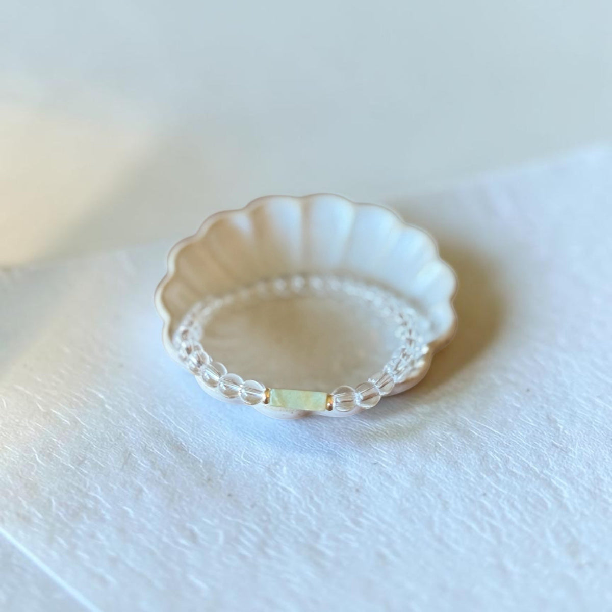 A delicate beaded bracelet with a scalloped cream-colored outer band and a thin inner band of clear beads, featuring a small pale green rectangular accent, displayed on a textured white surface.