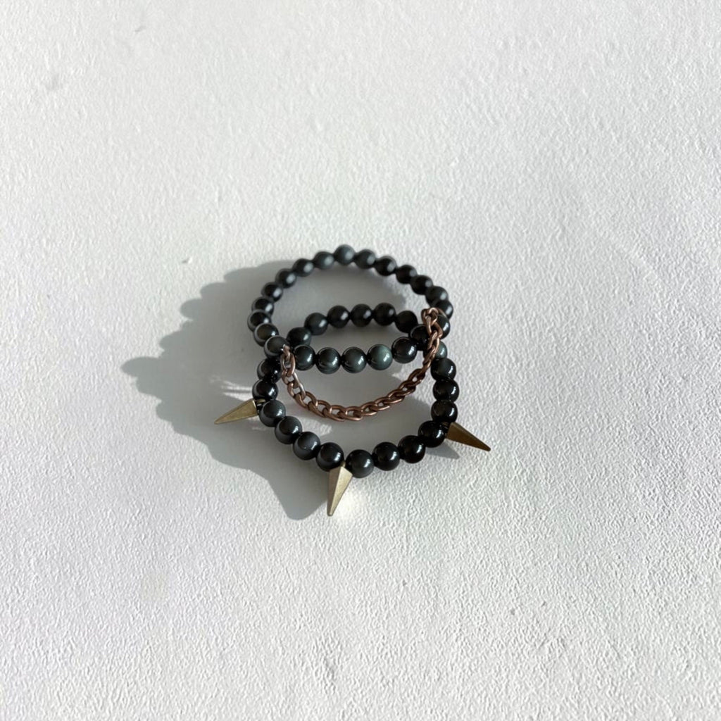 Two beaded rings stacked together on a textured white surface. The top and bottom rings are made of dark, polished black beads, while the center ring is a thin copper chain.