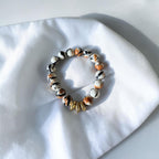 A beaded stretch bracelet featuring marbled white, black, and orange beads, accented by a cluster of small gold spacer beads, displayed on a white fabric surface.