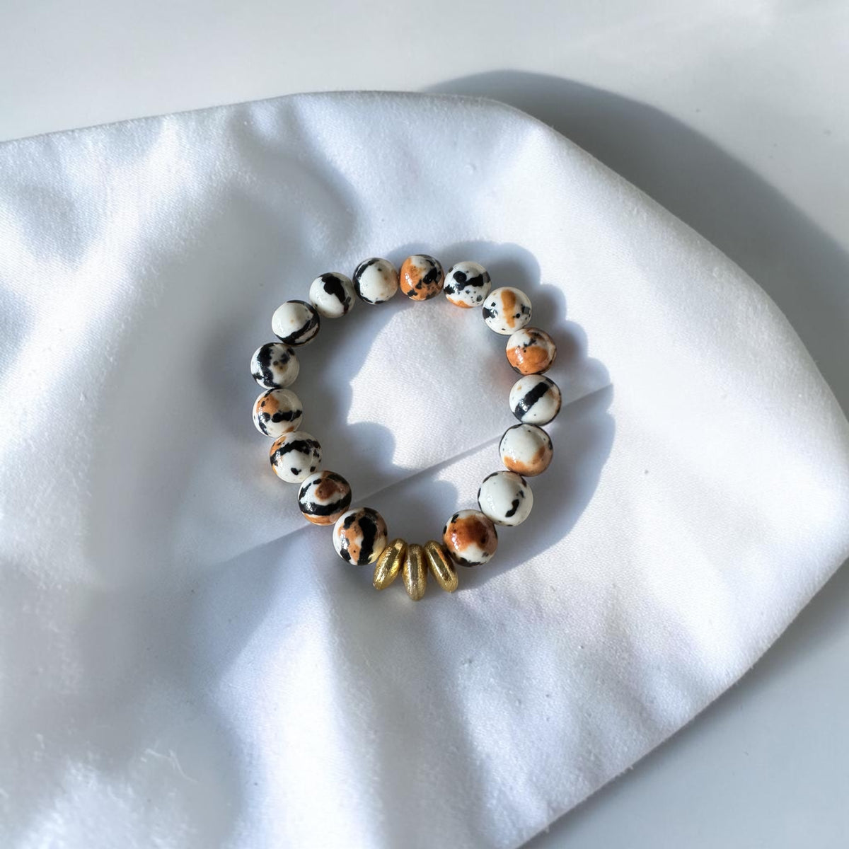 A beaded stretch bracelet featuring marbled white, black, and orange beads, accented by a cluster of small gold spacer beads, displayed on a white fabric surface.