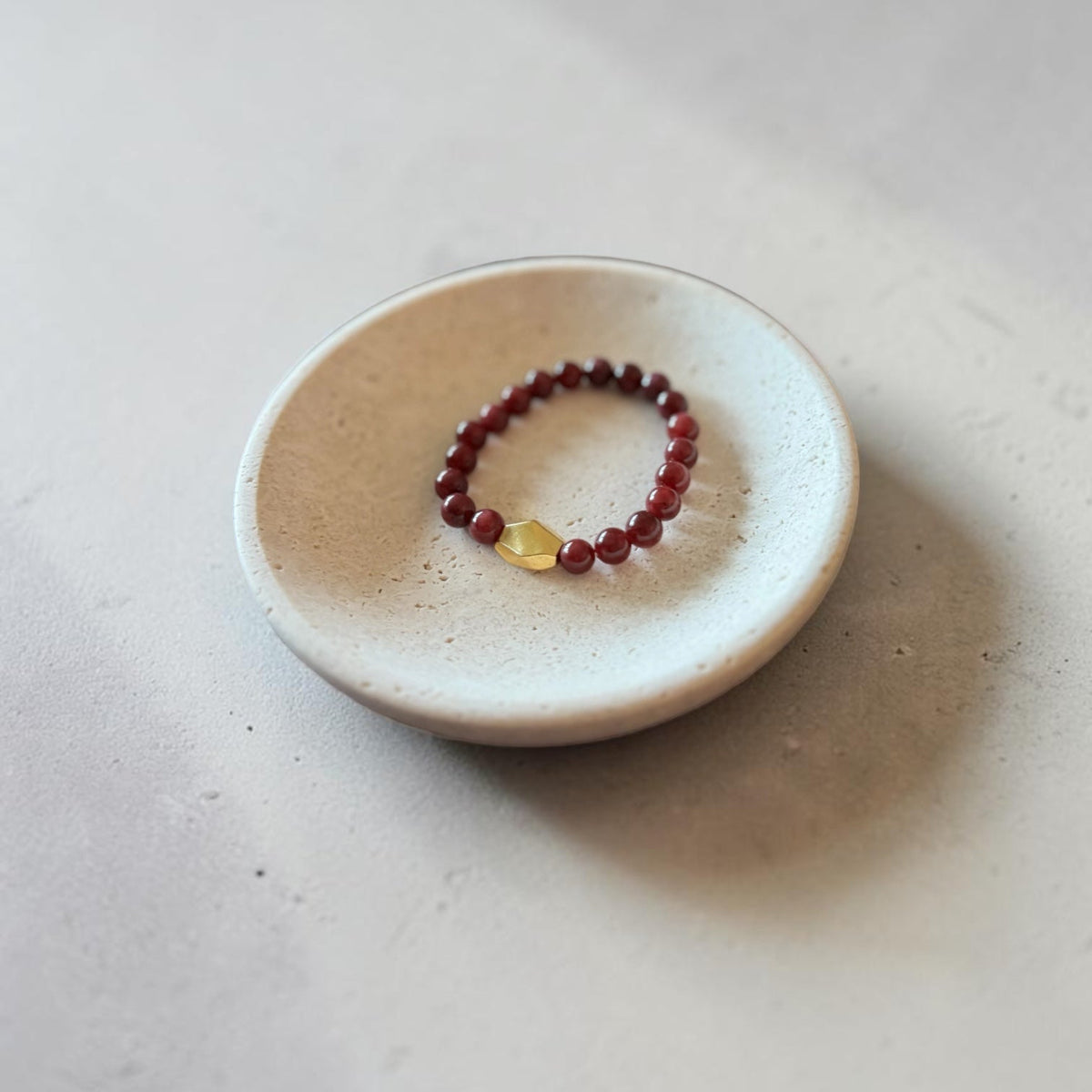 A beaded stretch bracelet with dark red beads and a small yellow accent stone, displayed on a small round ceramic dish against a neutral light gray background.