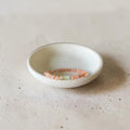 A delicate beaded bracelet in soft peach-pink and pale green tones rests inside a small, shallow white ceramic dish on a textured beige surface.