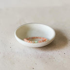 A delicate beaded bracelet in soft peach-pink and pale green tones rests inside a small, shallow white ceramic dish on a textured beige surface.